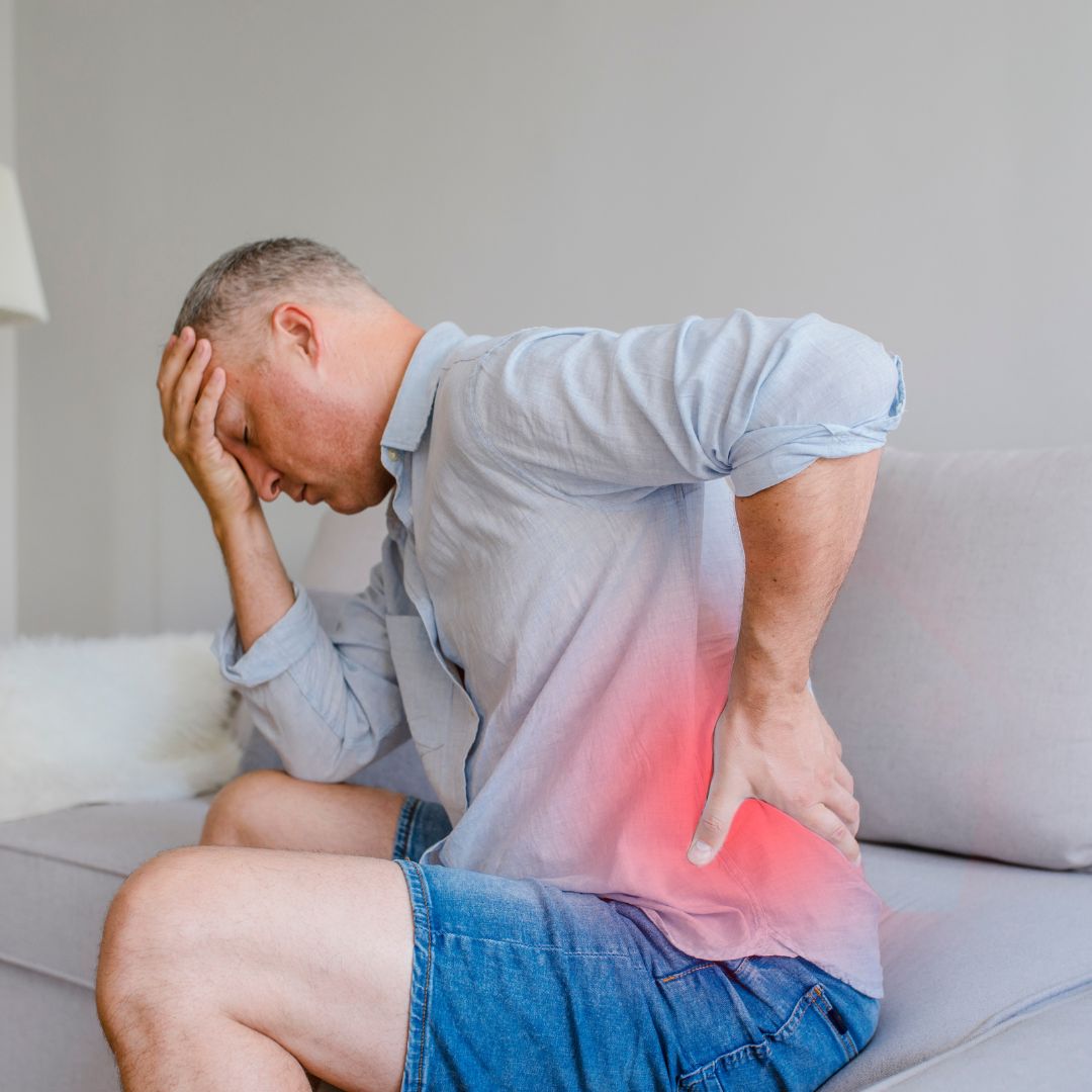 A man sits on a sofa, holding his back with one hand and his forehead with the other, indicating back pain. A red glow highlights the area of discomfort.