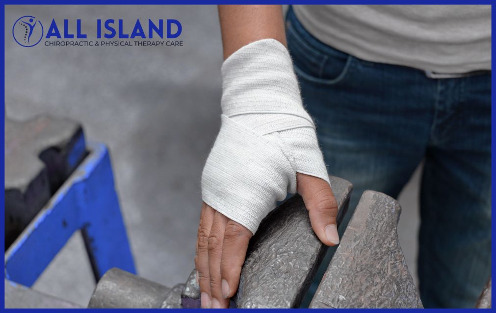 A person with a bandaged hand rests their hand on a metal surface; the All Island Chiropractic & Physical Therapy Care logo and Workers' Comp Doctor in Bayshore information are visible in the corner.