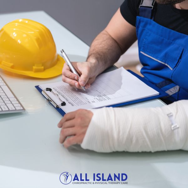 A person with a bandaged arm fills out a form on a clipboard at a desk with a computer keyboard and a yellow hard hat nearby.