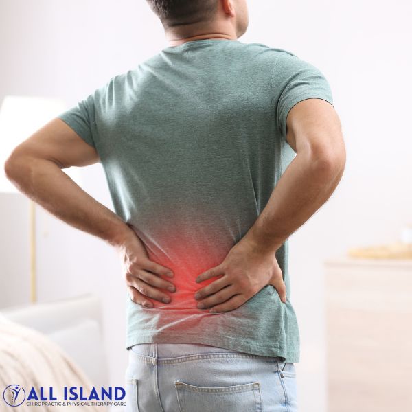 A man holding his lower back with both hands, indicating pain or discomfort, with a red highlight on his lower back. The "All Island" logo is visible in the corner, emphasizing Back Pain Treatment near Islip.