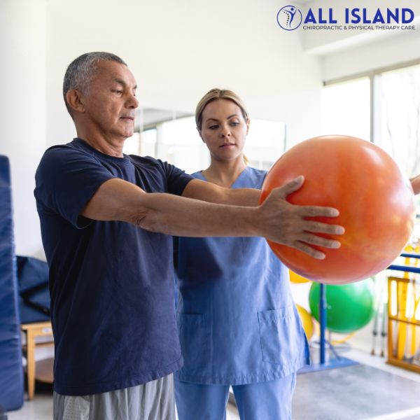 physical therapy islip An older man holds an exercise ball with both hands while a physical therapist observes and assists at a Physical Therapy near Islip clinic