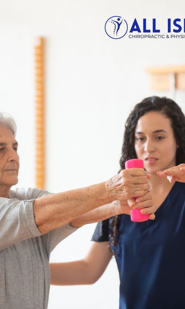An elderly woman exercises with a pink dumbbell under the guidance of a physical therapist at All Island Chiropractic & Physical Therapy Care 
