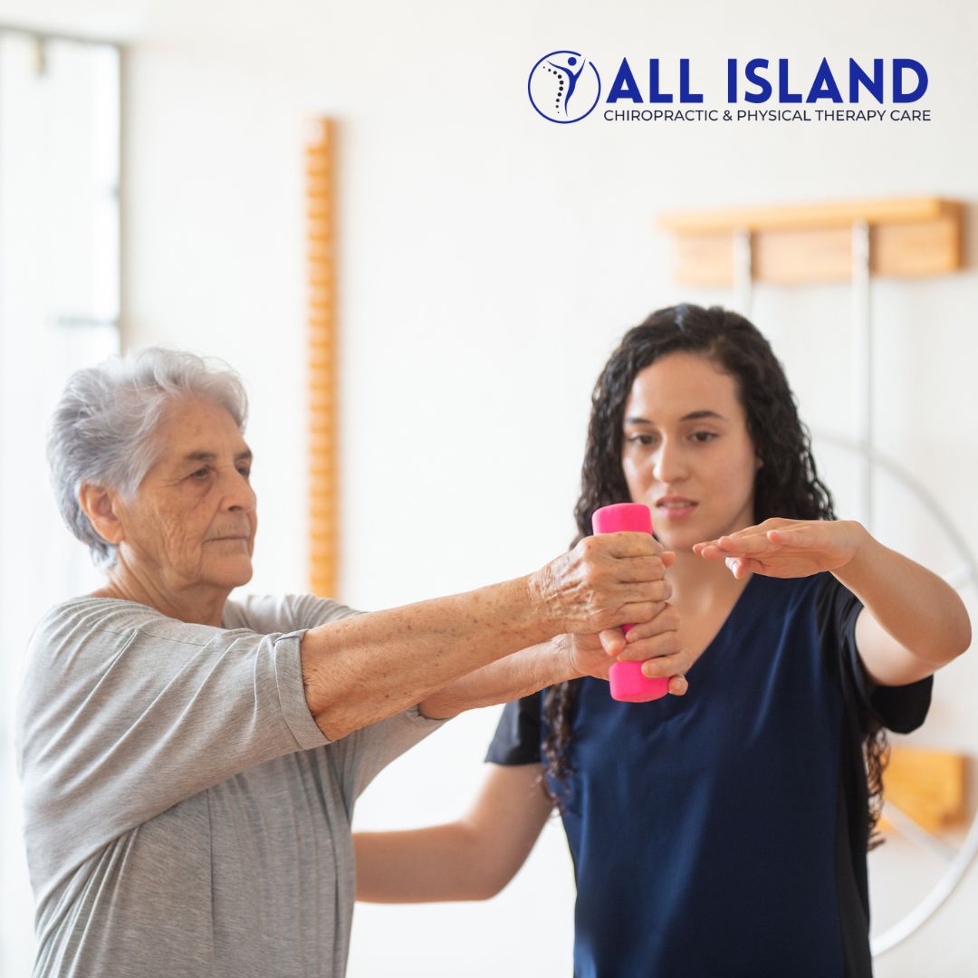 An elderly woman exercises with a pink dumbbell under the guidance of a physical therapist at All Island Chiropractic & Physical Therapy Care